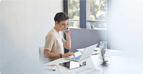 Eine Frau mit kurzen Haaren sitzt am Schreibtisch. Sie spricht in ihr Smartphone und hat einen Laptop sowie ein Tablet vor sich.